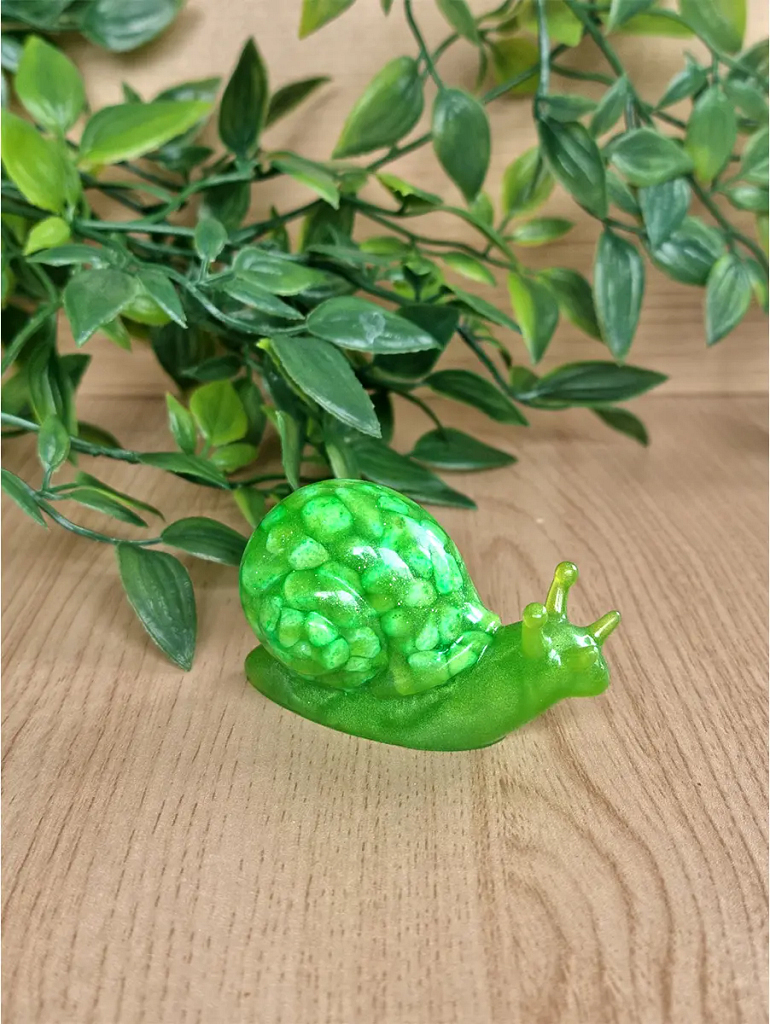 A small, translucent green resin snail sculpture with a shell resembling clustered gemstones, placed on a wooden surface amongst lush green foliage.