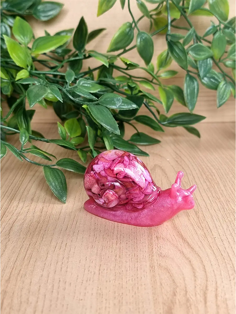 A vibrant pink resin snail sculpture featuring a shell filled with crushed pink flower petals and glitter, resting on a wooden surface.