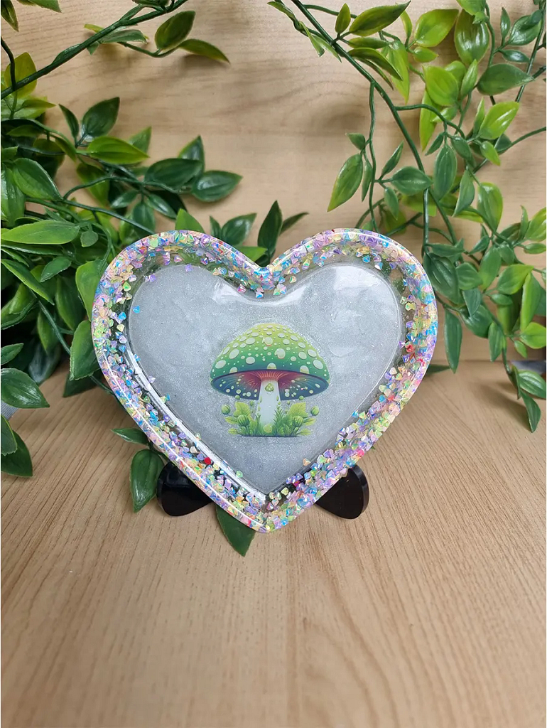 Handcrafted resin art dish in a heart shape, featuring a whimsical mushroom motif surrounded by shimmering glitter and natural greenery.