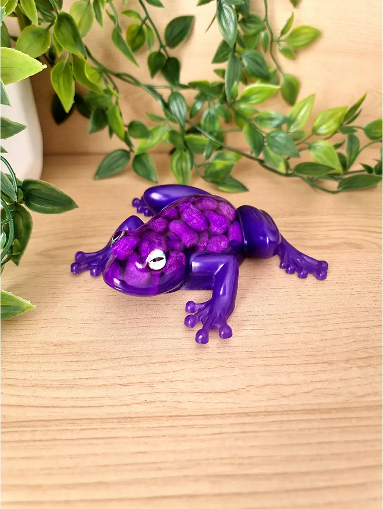 A whimsical purple resin frog sculpture with purple pebbles resting on a wooden surface with lush green foliage in the background.