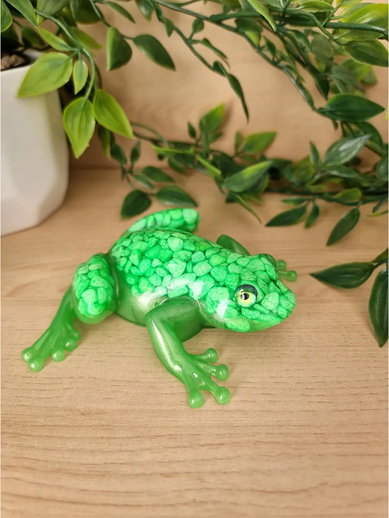 A vibrant emerald green textured resin frog figurine with green pebbles, placed on a light wood surface with artificial greenery in the background.