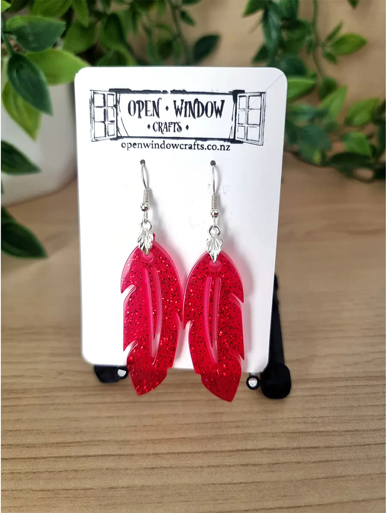 Sparkling red feather-shaped resin earrings dangle from silver hooks on a branded card.