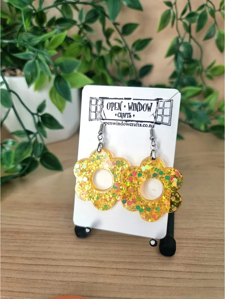 Golden flower-shaped resin earrings with glitter and colourful flecks, suspended from silver hooks.