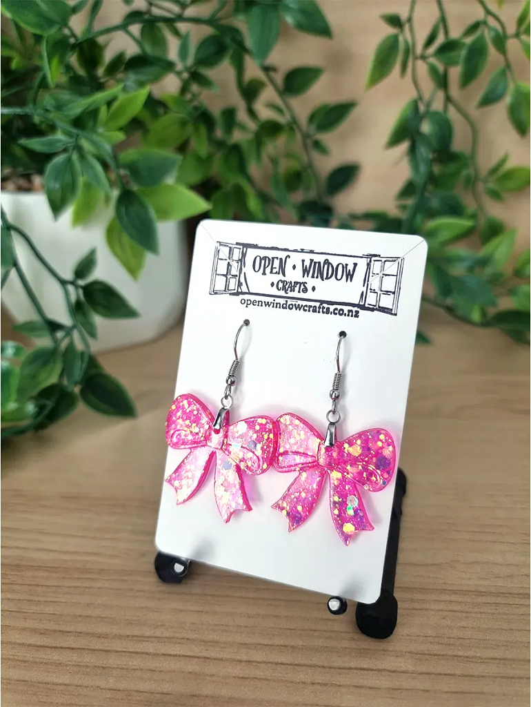 Pink glitter bow resin earrings dangle from silver hooks on a branded card.
