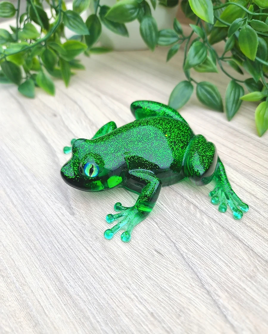 A vibrant emerald green resin frog figurine adorned with glittering particles, featuring striking blue and green eyes.