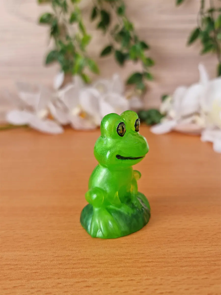 A small, translucent green resin figurine of a frog sits on a green leaf base, featuring yellow-rimmed eyes and a subtle smile.