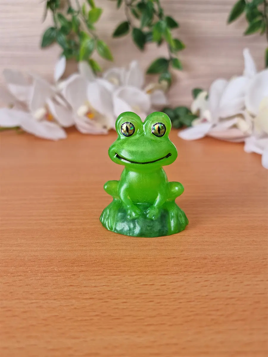 A vibrant green, translucent resin frog figurine with shimmering, multi-faceted eyes sits on a textured green base.