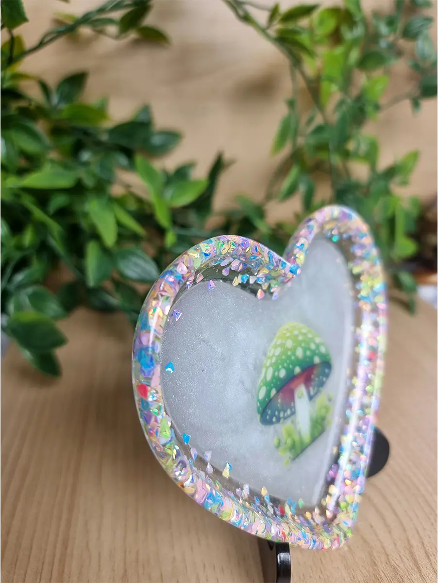 A heart-shaped resin art piece featuring a whimsical green mushroom illustration surrounded by iridescent glitter and set against a soft, luminous background.