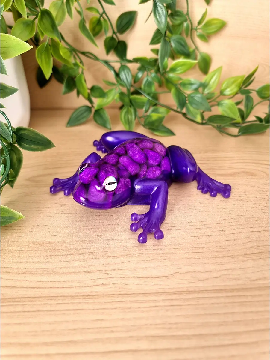 A whimsical purple resin frog sculpture with purple pebbles resting on a wooden surface with lush green foliage in the background.
