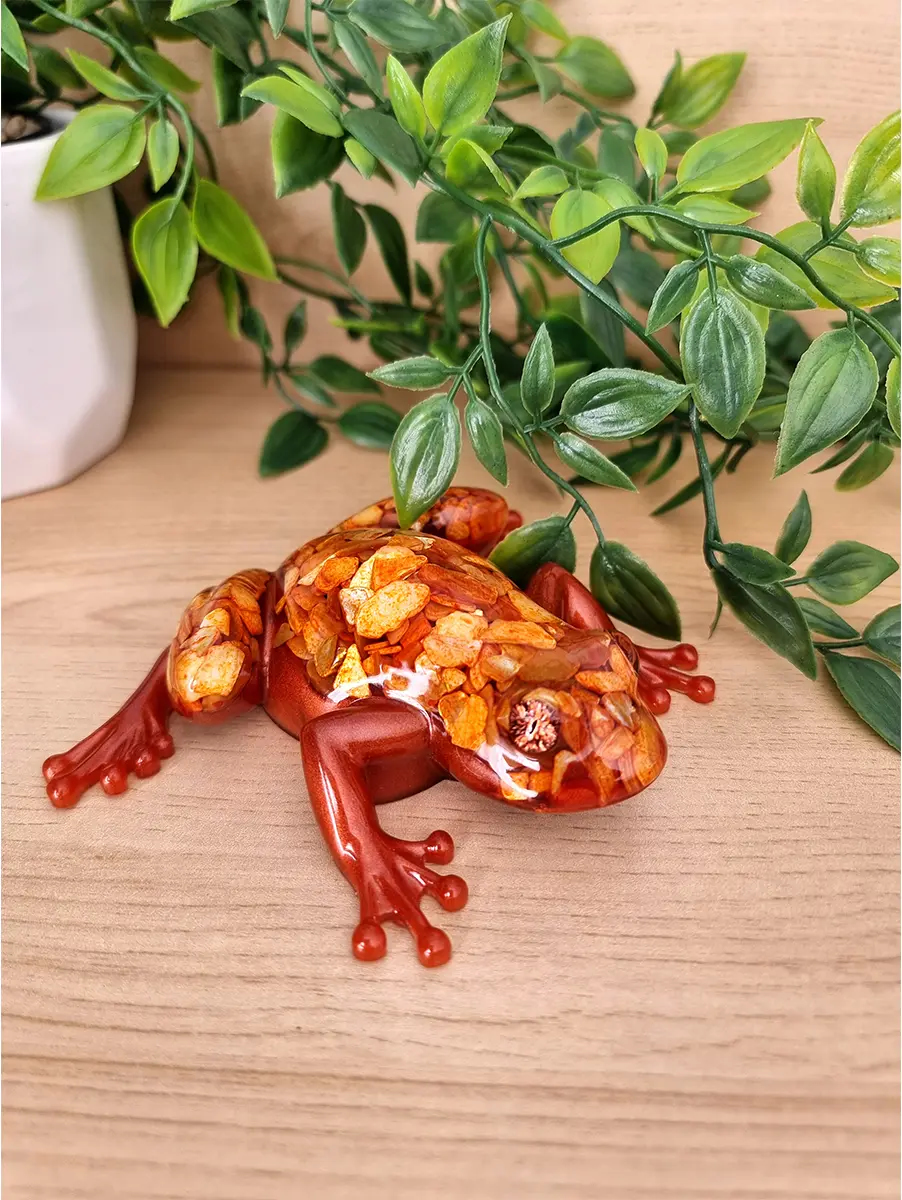 A detailed resin sculpture of a frog, with iridescent metallic red limbs and a body filled with shimmering amber-like crushed shells.