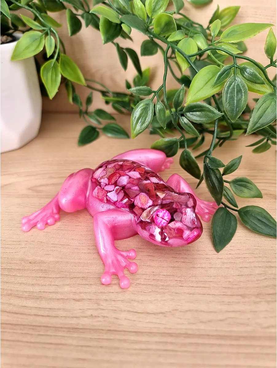 A vibrant pink resin frog sculpture features a shimmering geode-style back filled with crushed pink and purple crushed shells, resting on a wooden surface.