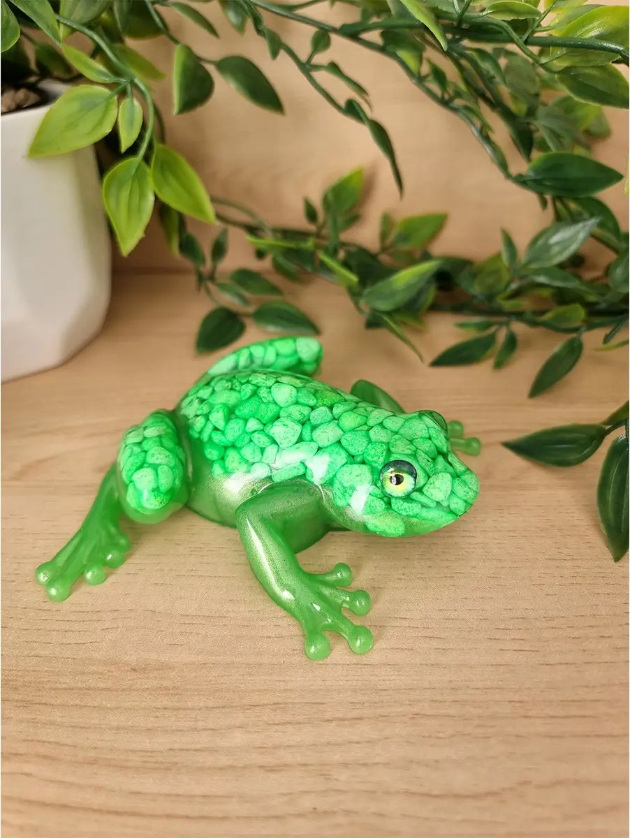 A vibrant emerald green textured resin frog figurine with green pebbles, placed on a light wood surface with artificial greenery in the background.