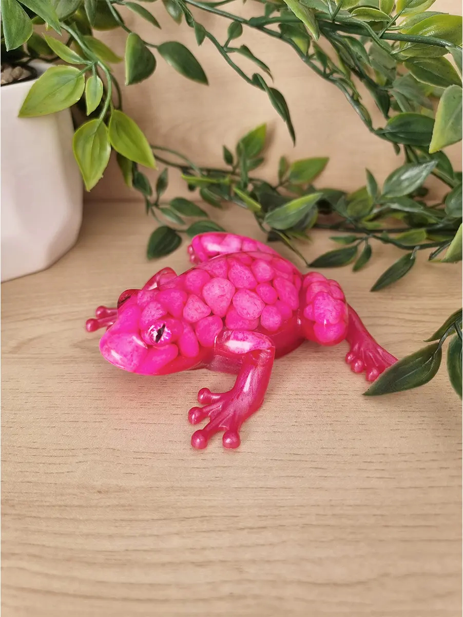 A brightly coloured pink resin sculpture of a frog, with a textured back resembling small pebbles and translucent limbs.