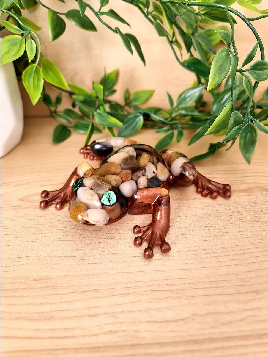 A detailed handcrafted resin frog figurine filled with smooth pebbles and featuring striking green gem eyes and copper-toned legs.