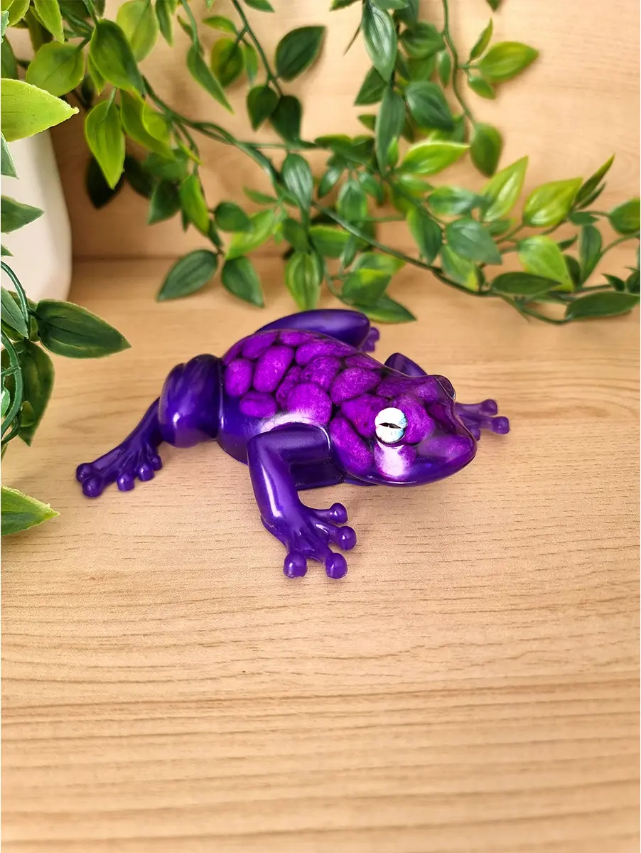 A glossy royal purple resin frog figurine with purple stones and white eyes rests on a light wood surface.