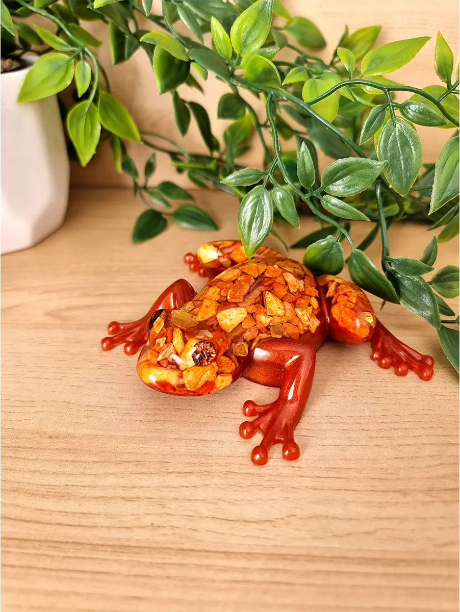 A handcrafted resin sculpture of a frog with a glossy orange body and a back adorned with small amber-colored crushed shells and a faceted eye.