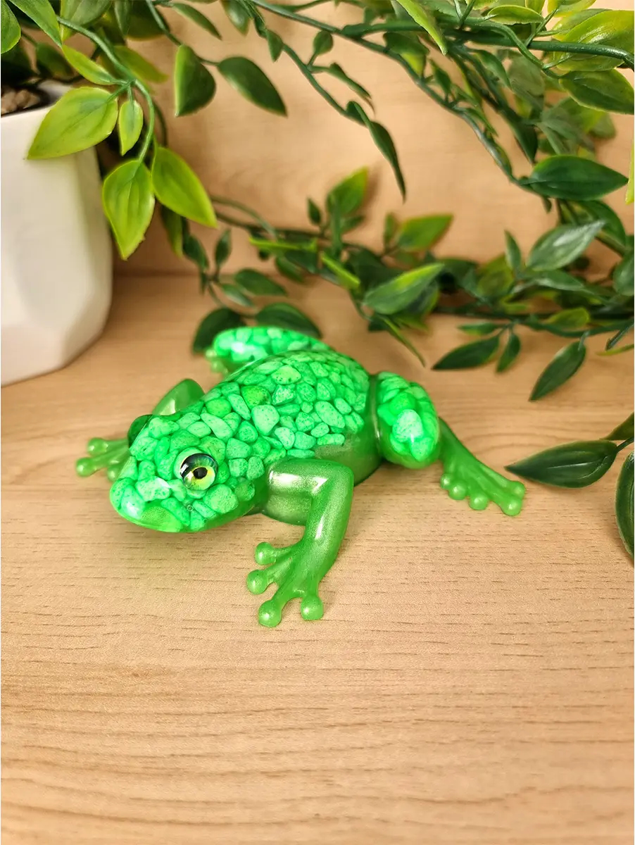 A vibrantly colored emerald green frog sculpture with a textured back and shiny finish rests on a wooden surface, surrounded by lush green foliage.