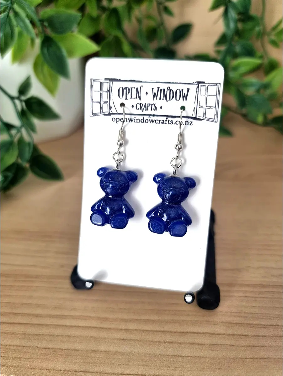 Pair of deep blue, glossy resin gummy teddy bear charm earrings dangle from silver hooks, presented on a white Open Window Crafts branded display card.