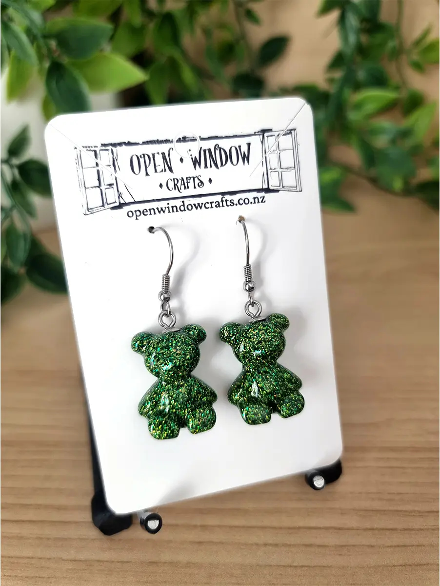 Pair of sparkling emerald green gummy bear resin earrings dangling from silver hooks, presented on a branded card.