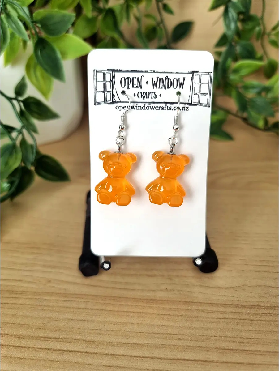 Handcrafted orange gummy bear shaped resin earrings dangle from silver hooks, presented on an Open Window Crafts display card.