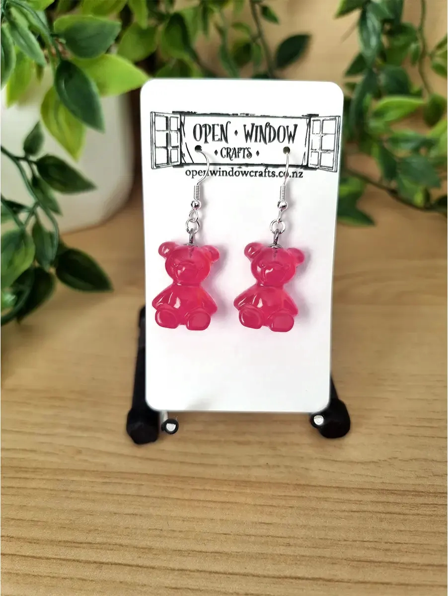 Vibrant raspberry-pink gummy bear resin dangle earrings hang from silver hooks on a white display card, showcasing their playful, cute design.