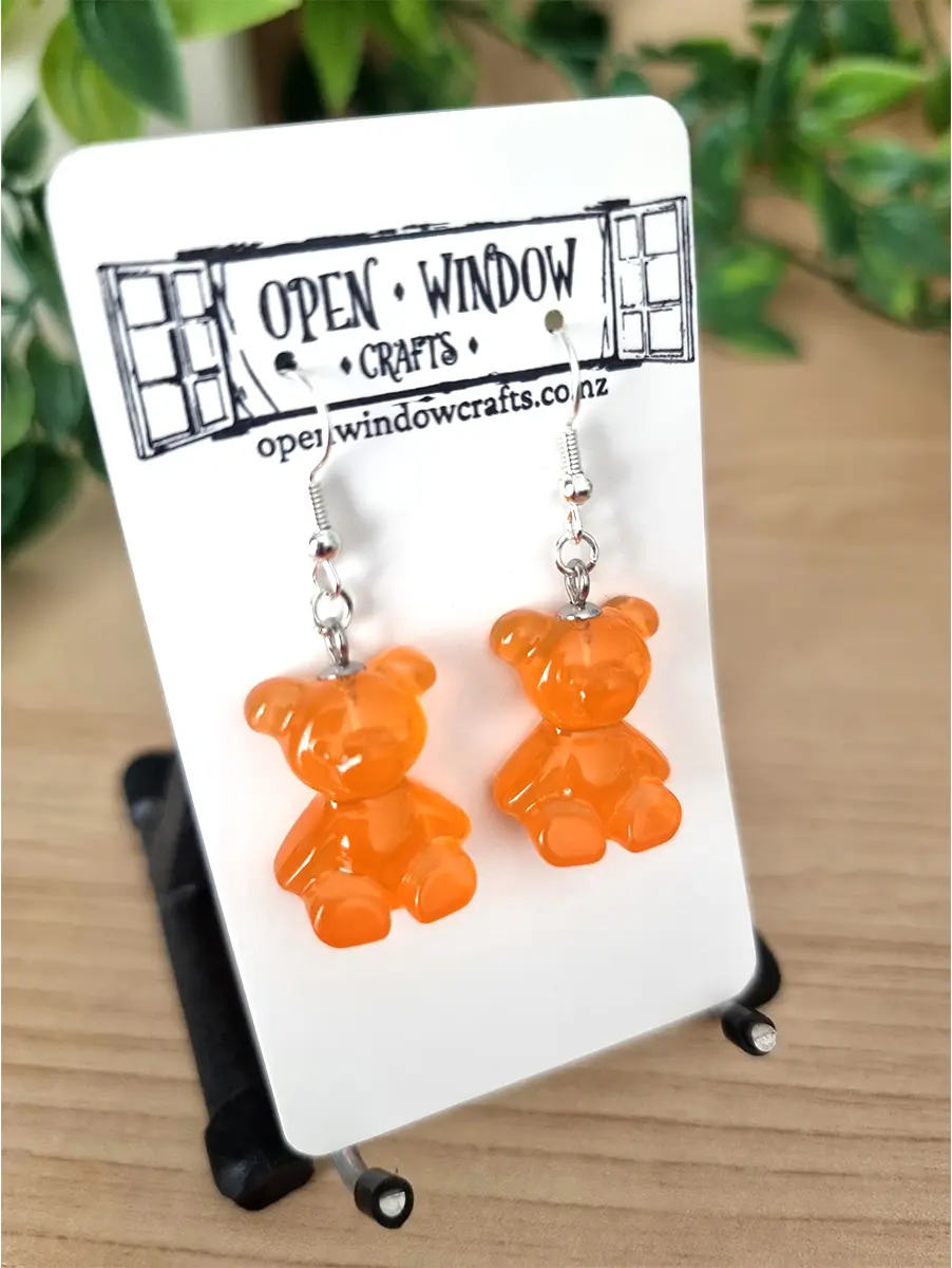 Two translucent orange gummy bear resin dangle earrings are showcased on a white Open Window Crafts card, each suspended from a silver fishhook.
