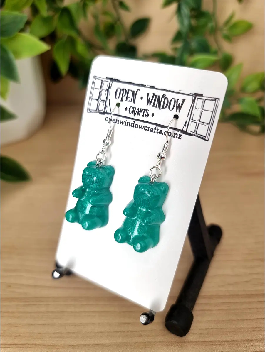 Teal gummy bear resin earrings hang from silver hooks on a white display card.