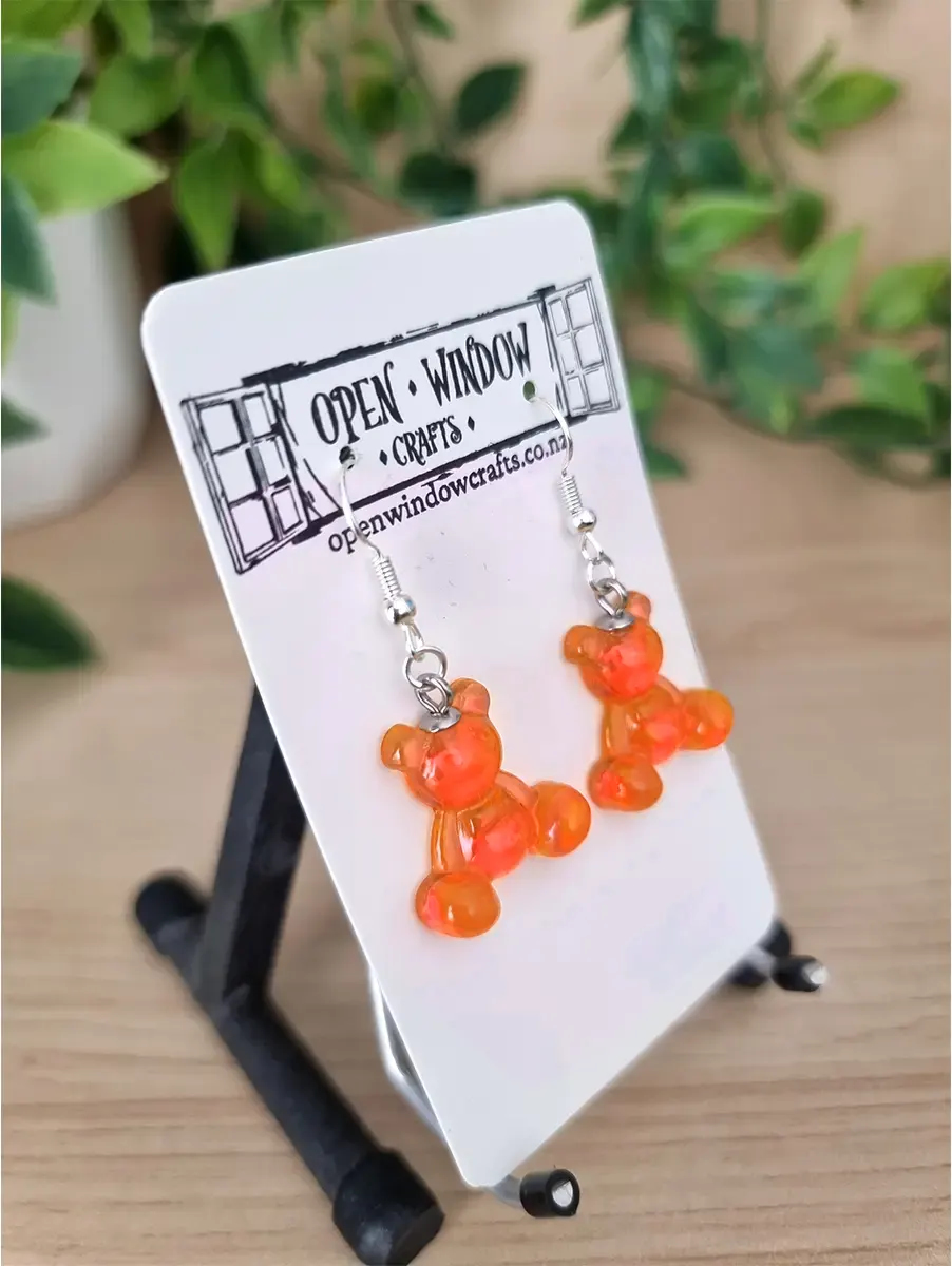 Translucent orange gummy bear earrings dangle from silver hooks on a white logoed card.