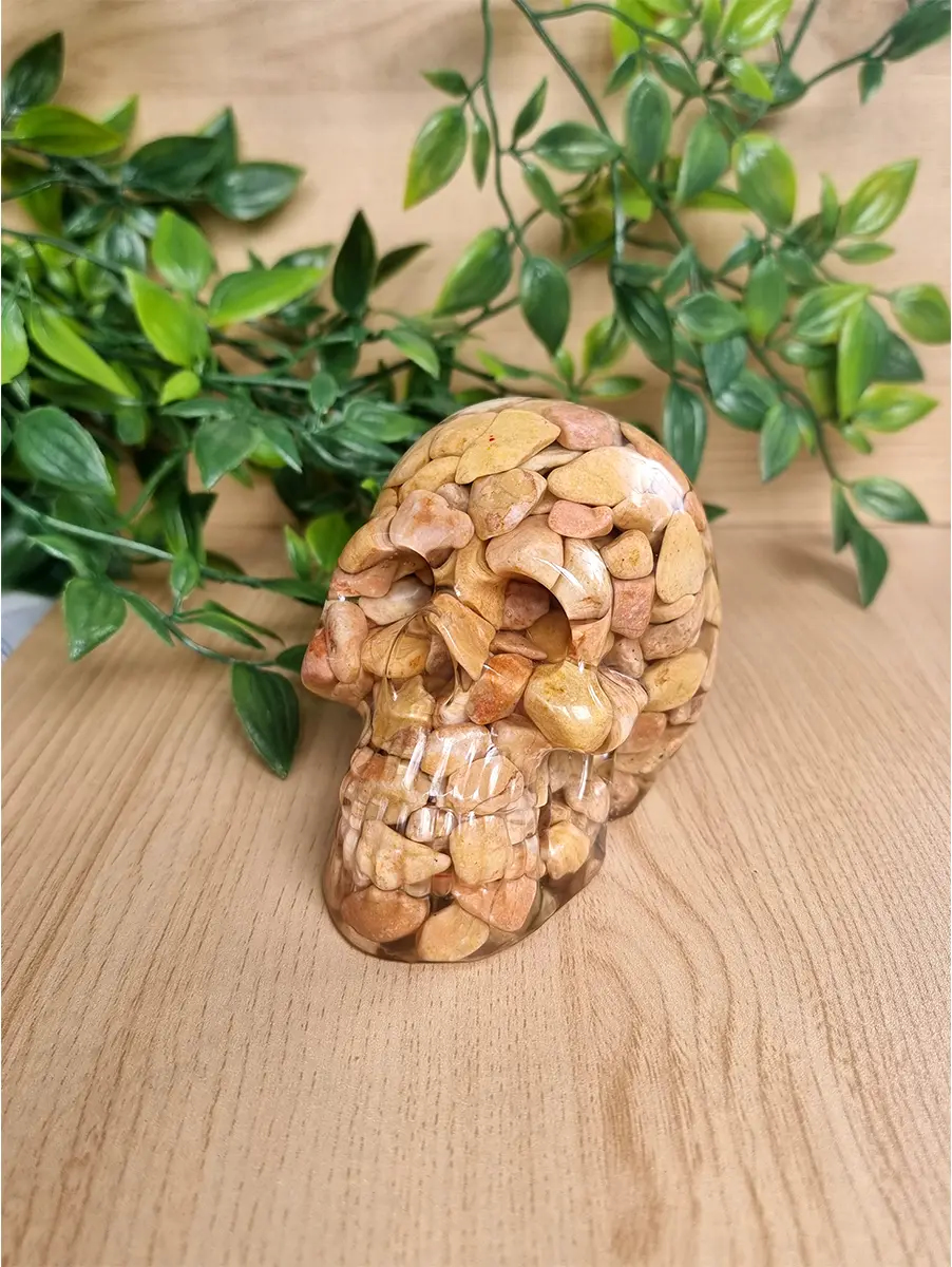 Polished resin encapsulates natural river stones forming a unique skull sculpture, casting subtle shadows on a wooden surface with green foliage.