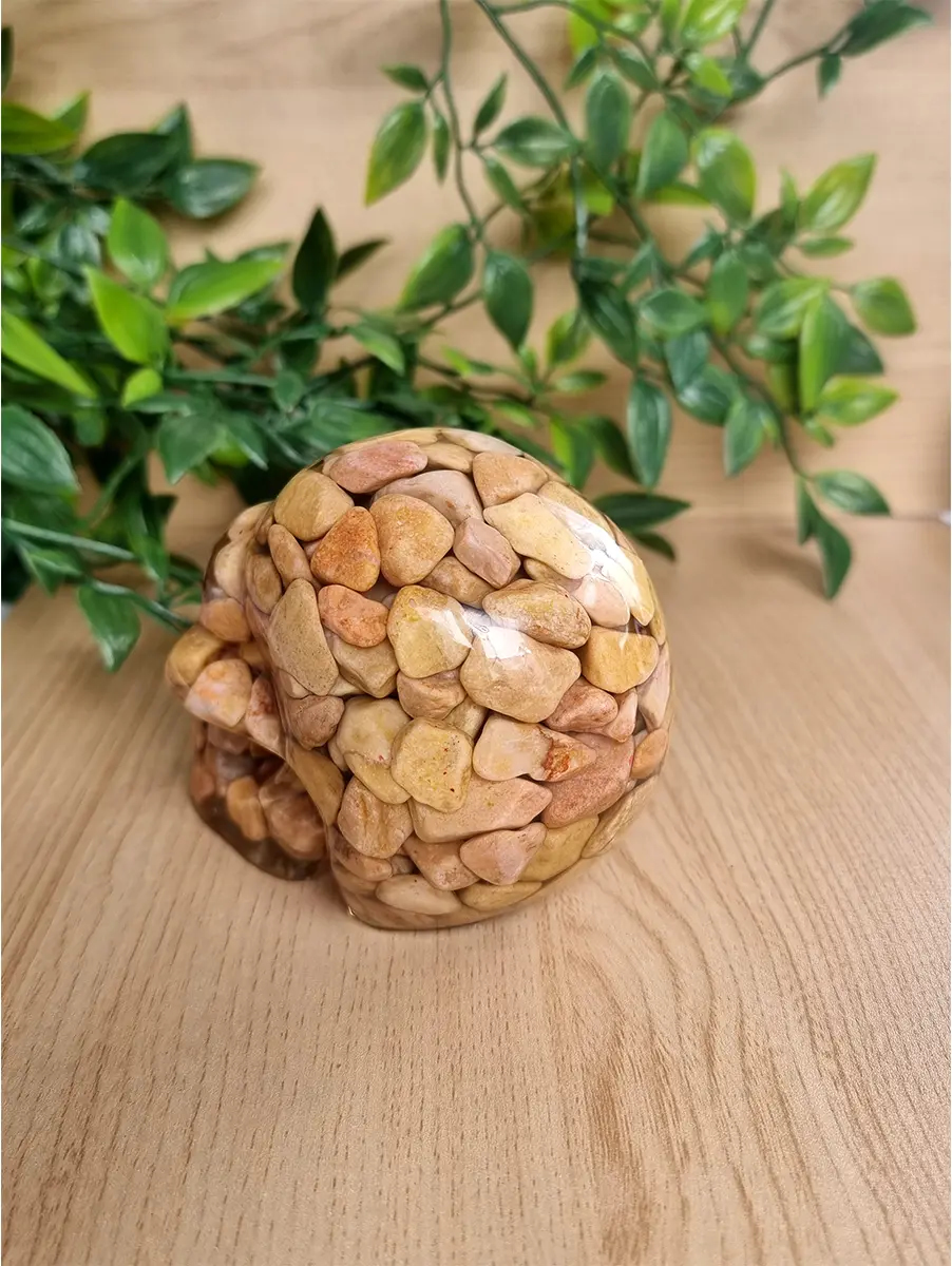 Pebble-filled resin skull sculpture sits on a wooden surface, adorned by greenery.