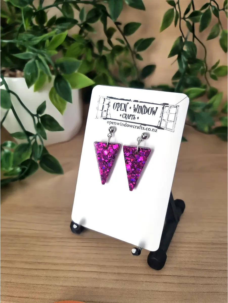 Fuchsia glitter triangle resin earrings sparkle on a white display card, suspended by silver hooks.