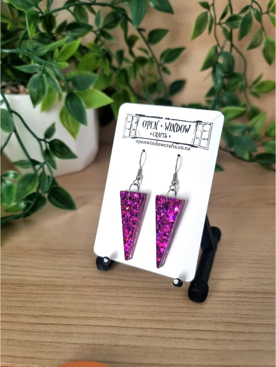 Sparkling pink triangle resin earrings dangle from silver hooks on a white branded card, showcasing iridescent glitter.