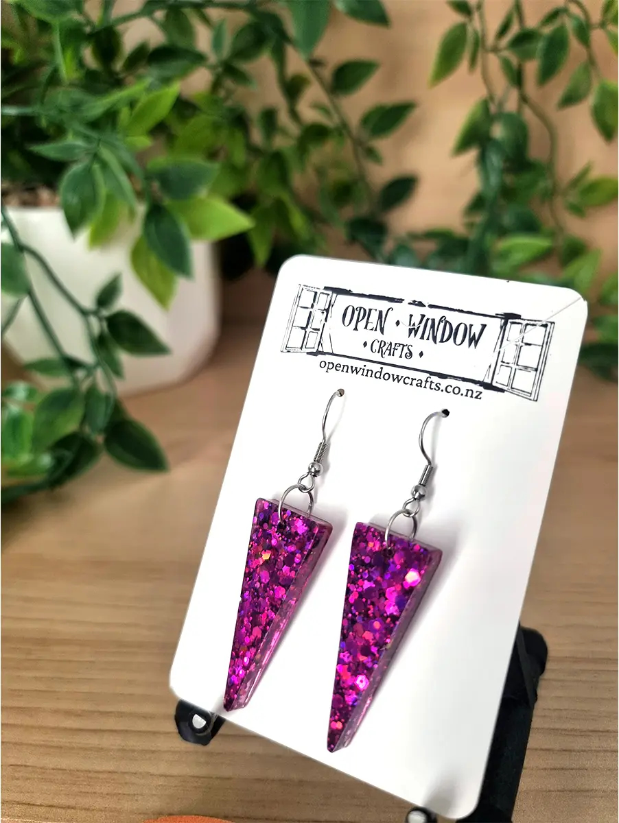 Two magenta resin triangle earrings dangle, filled with bright glitter and suspended on silver hooks.