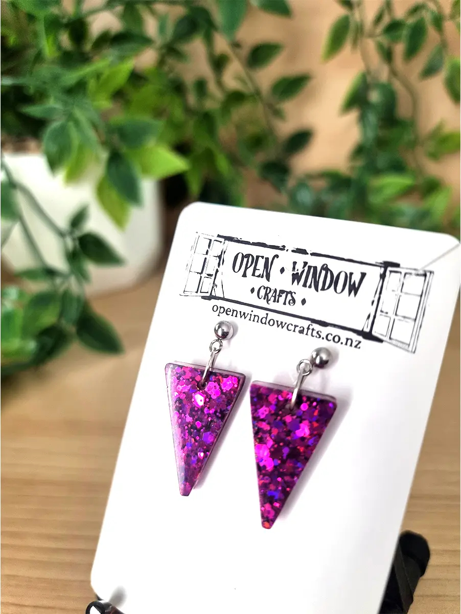 Triangular fuchsia glitter resin earrings dangle from silver posts, showcasing a vibrant, shimmering effect.