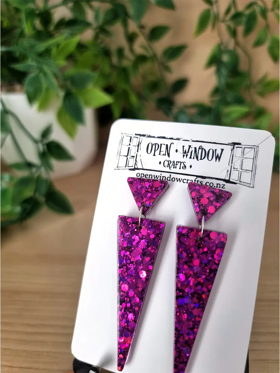Triangular pink glitter resin earrings dangle from silver hooks on a branded card.