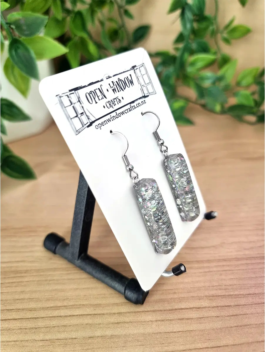 Two clear resin column earrings with iridescent glitter sparkles dangle from silver hooks on a white display card.