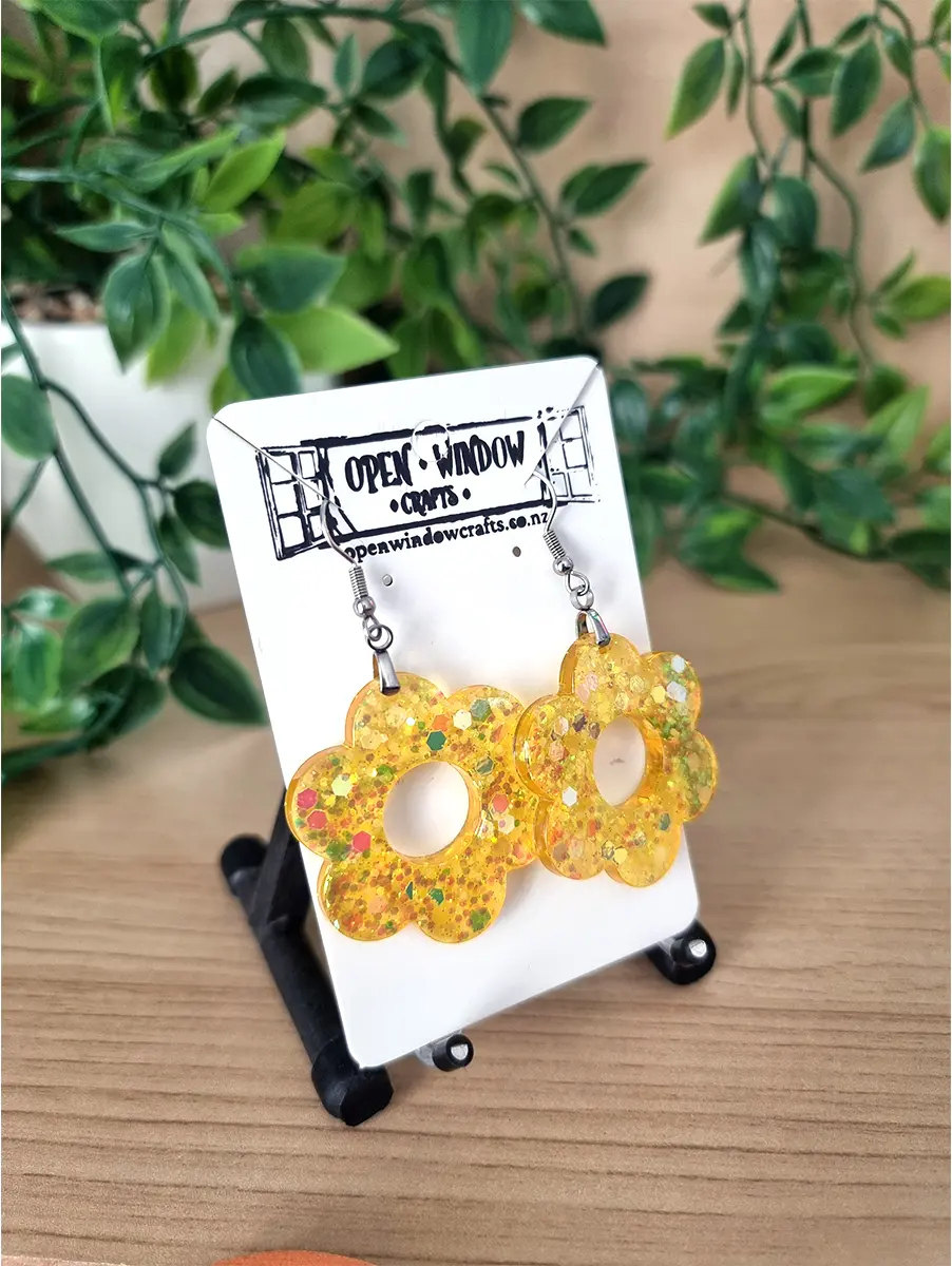 Yellow flower-shaped resin earrings glitter accents dangle from silver hooks on a white card.