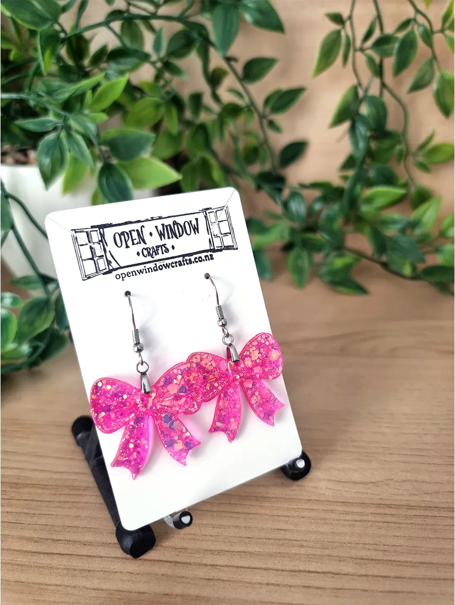 Pink glitter bow earrings dangle from silver hooks; handcrafted by Open Window Crafts.