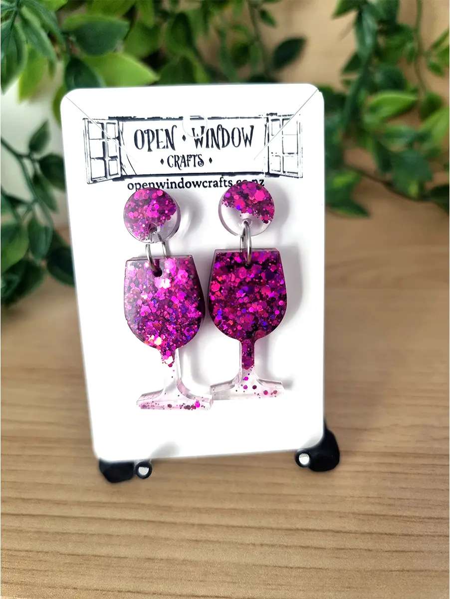 Sparkling raspberry wine glass earrings crafted from resin glitter hang from silver hooks on a branded card.